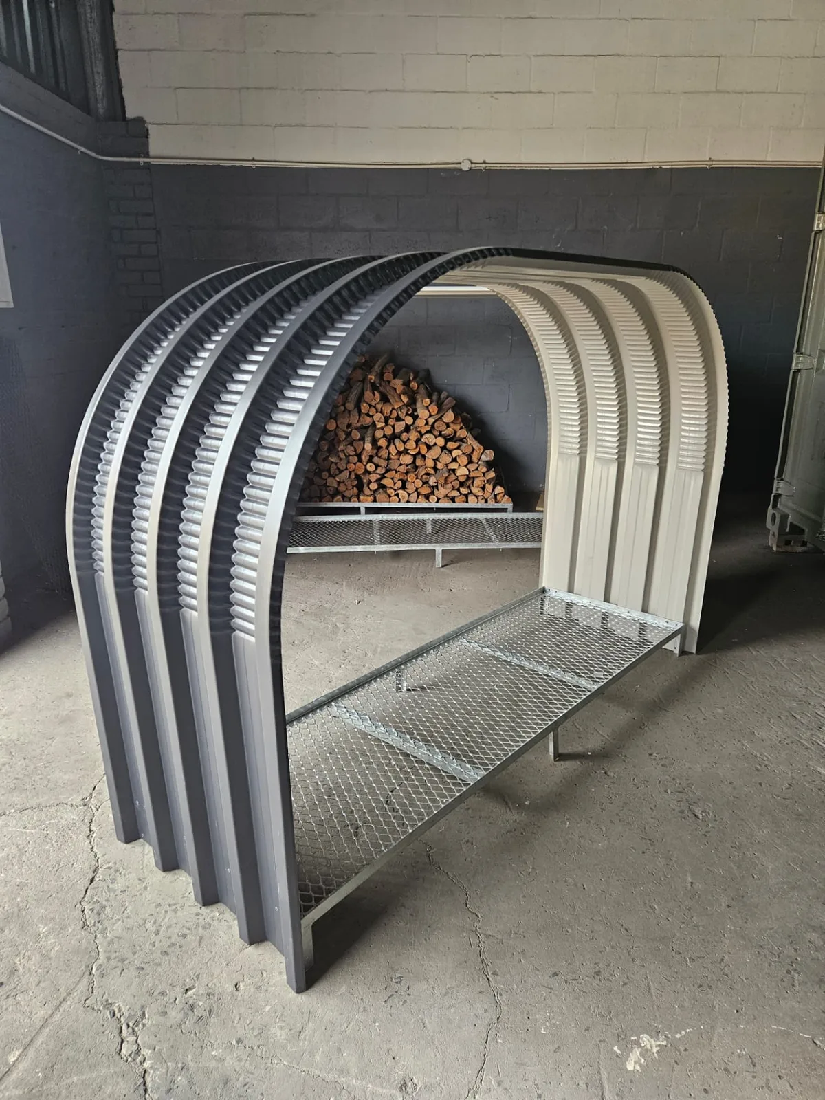 Firewood shed arch frame in charcoal and cream with a galvanised mesh base, stacked logs behind