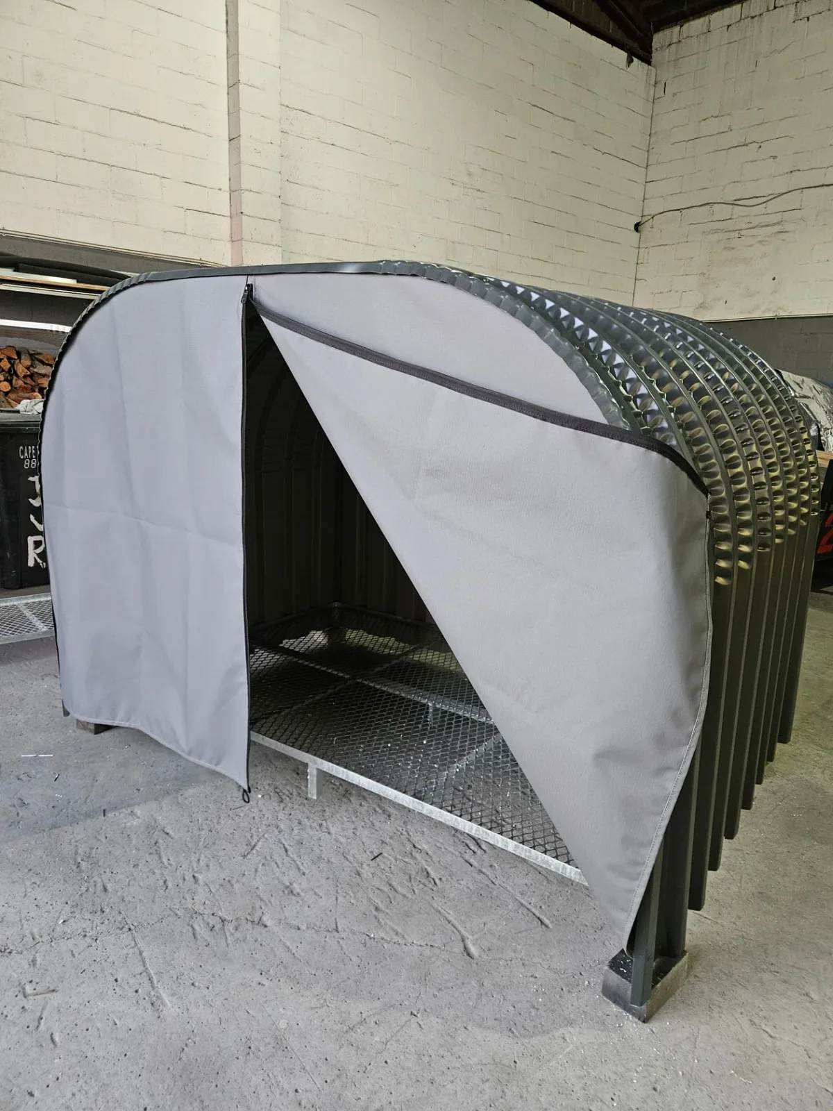 Firewood shed with grey canvas cover unzipped and folded back, showing the galvanised mesh base inside the workshop