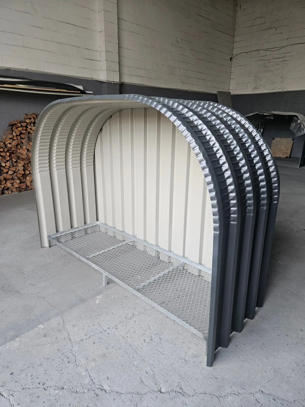 Firewood shed with charcoal curved corrugated arches, cream corrugated back panel and galvanised mesh base