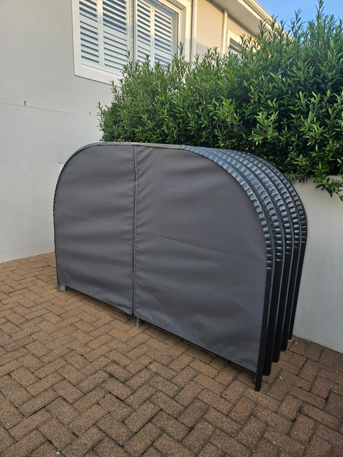 Charcoal firewood shed with a grey zipped canvas cover installed on brick paving beside a house wall