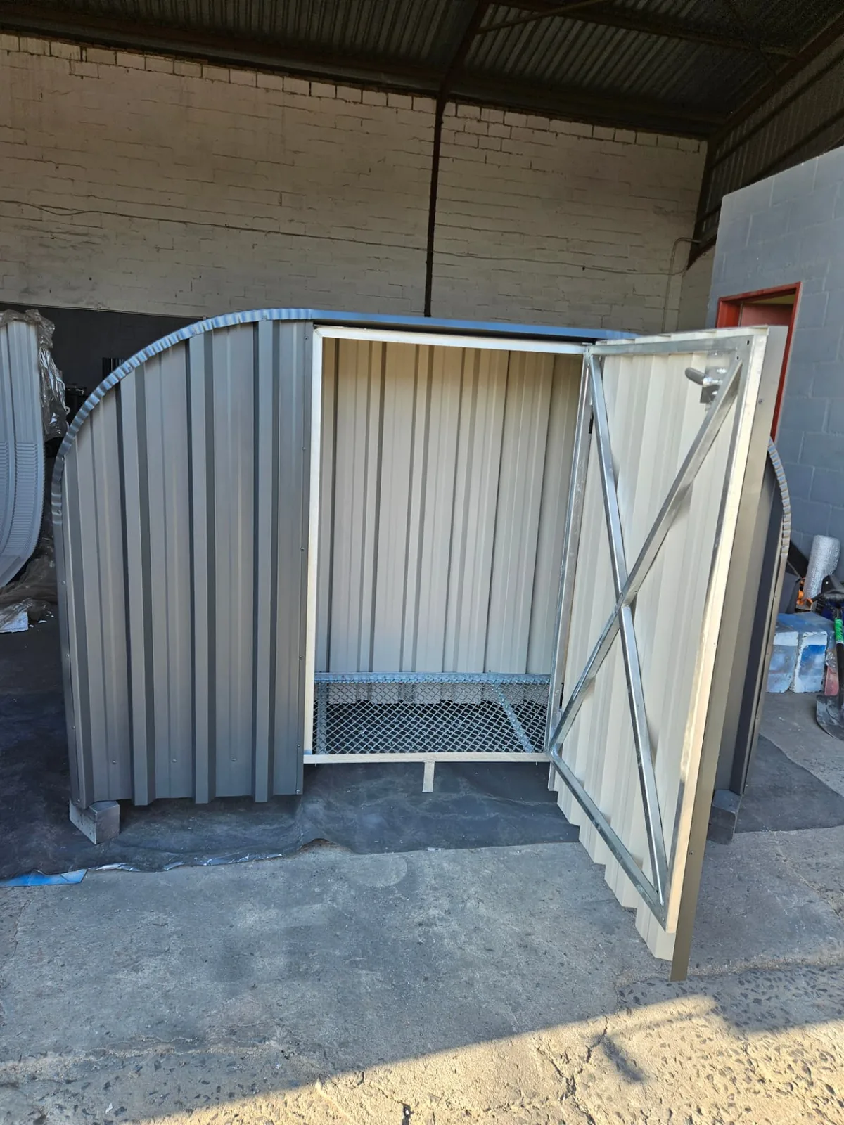 Firewood shed with the lockable steel door swung open, showing the braced door frame and galvanised mesh base