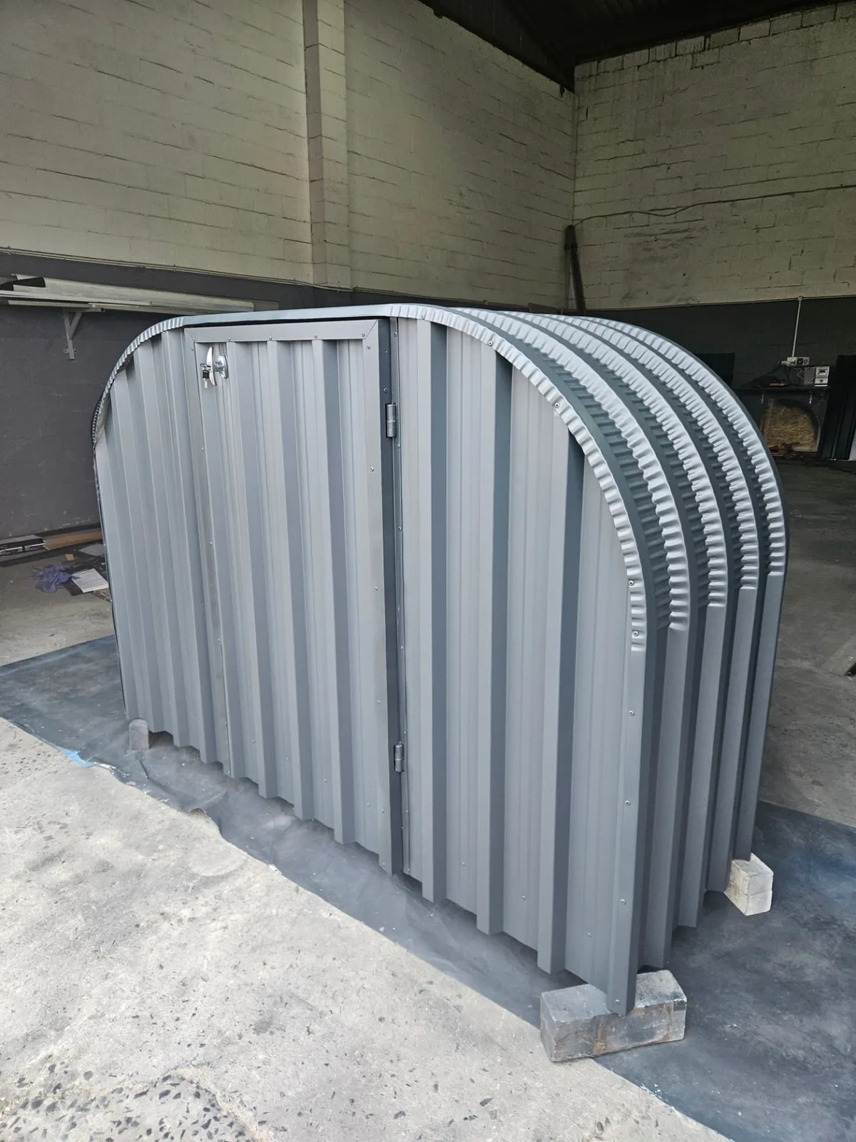 Grey firewood shed with a closed lockable door and curved corrugated roof arches in the workshop