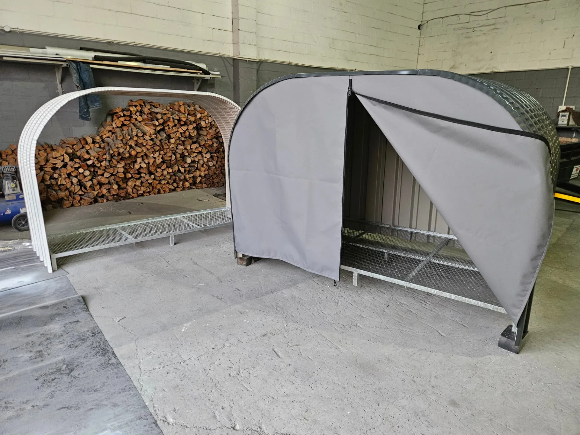 Two firewood sheds side by side in the workshop, one cream frame and one charcoal frame with a grey canvas cover