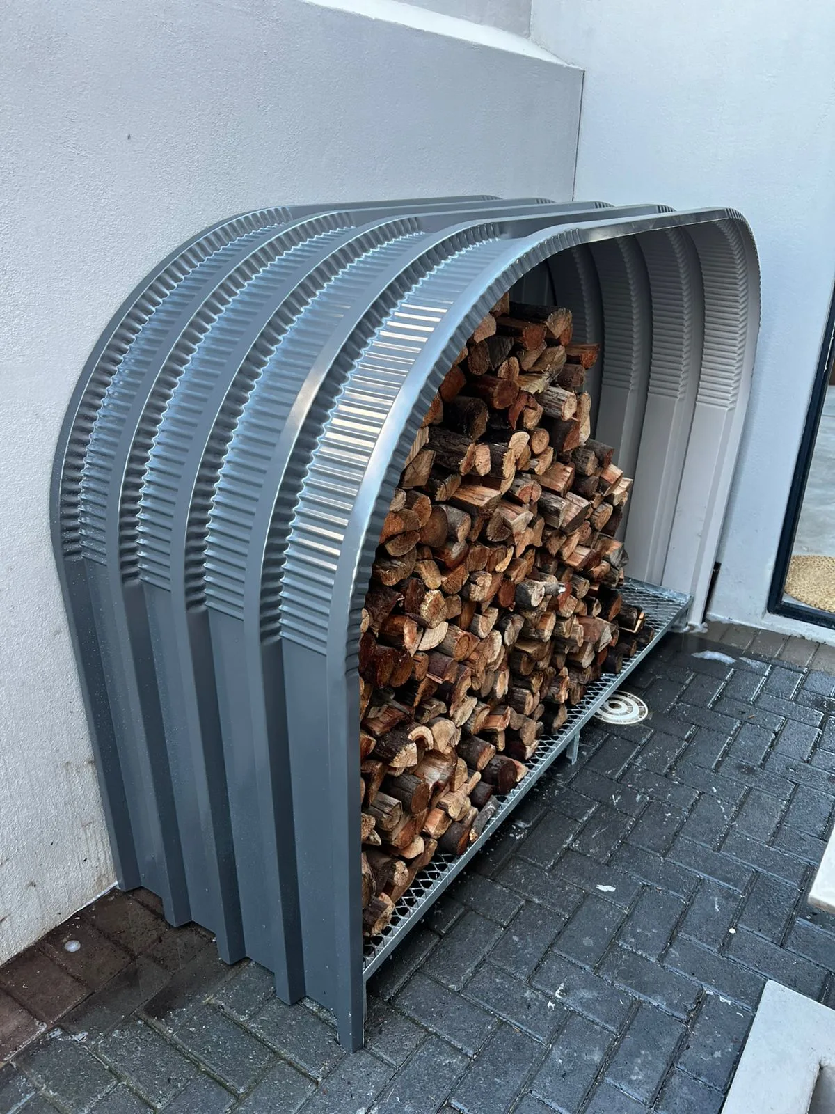 Charcoal firewood shed filled with stacked split logs against a white wall on brick paving