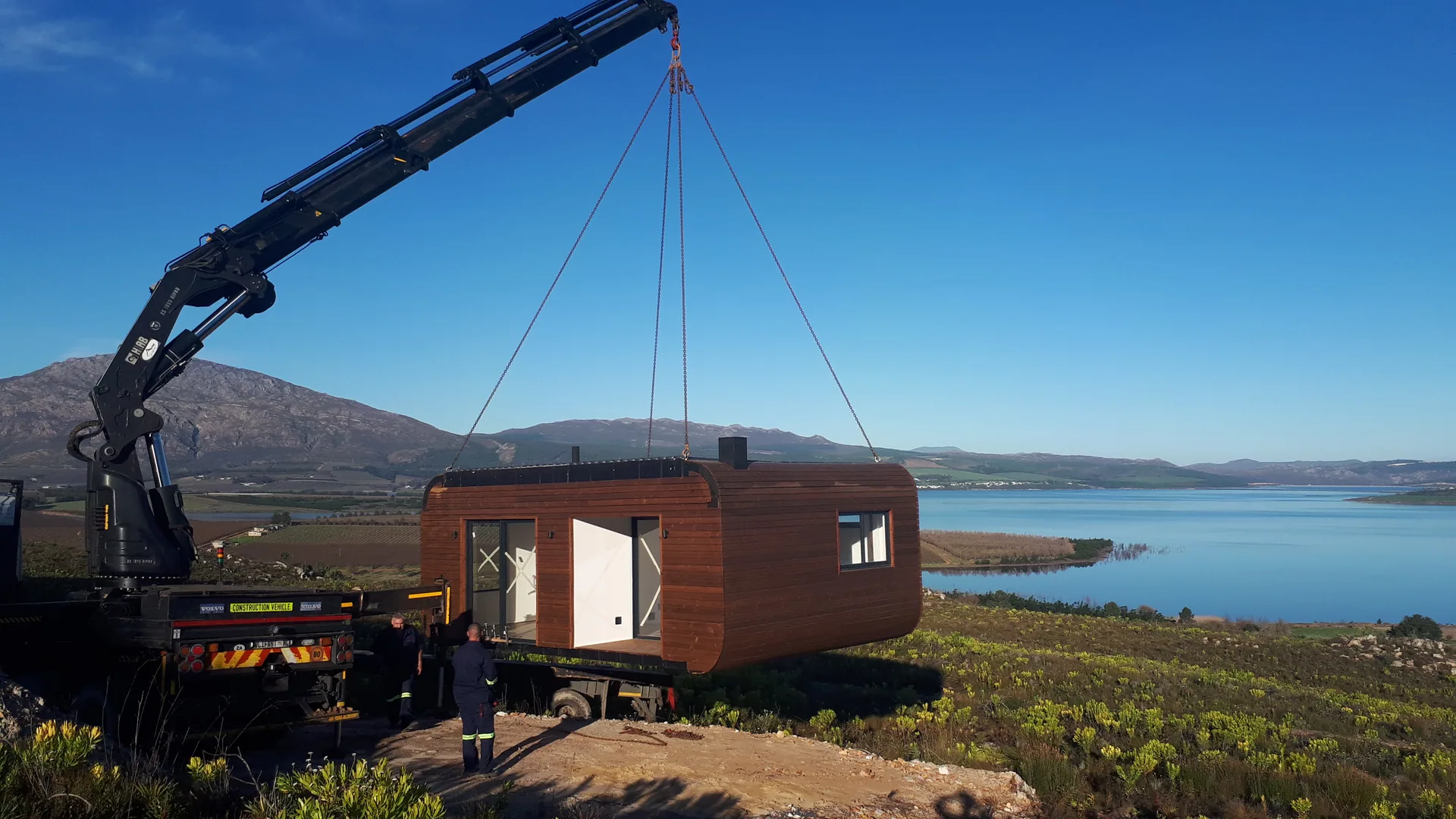 Crane lifting a dark timber-clad pod onto a hilltop site overlooking a lagoon and mountains