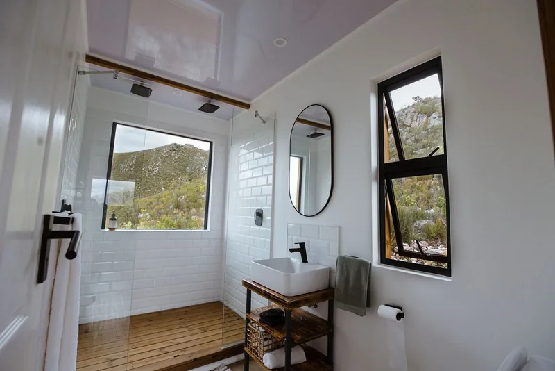 White-tiled container house bathroom with a timber vanity shelf, round mirror, black-framed window and walk-in shower