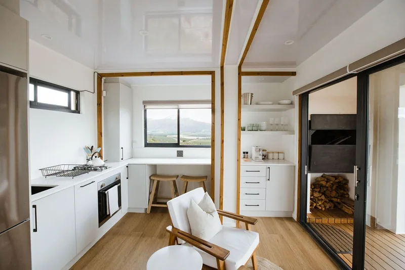 Open-plan container house interior with a white kitchen, oven, timber ceiling battens and a built-in sleeping alcove