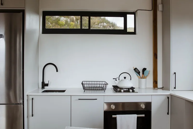 Fitted container house kitchen with white cabinets, black tap, gas hob, oven and stainless fridge beneath a horizontal window
