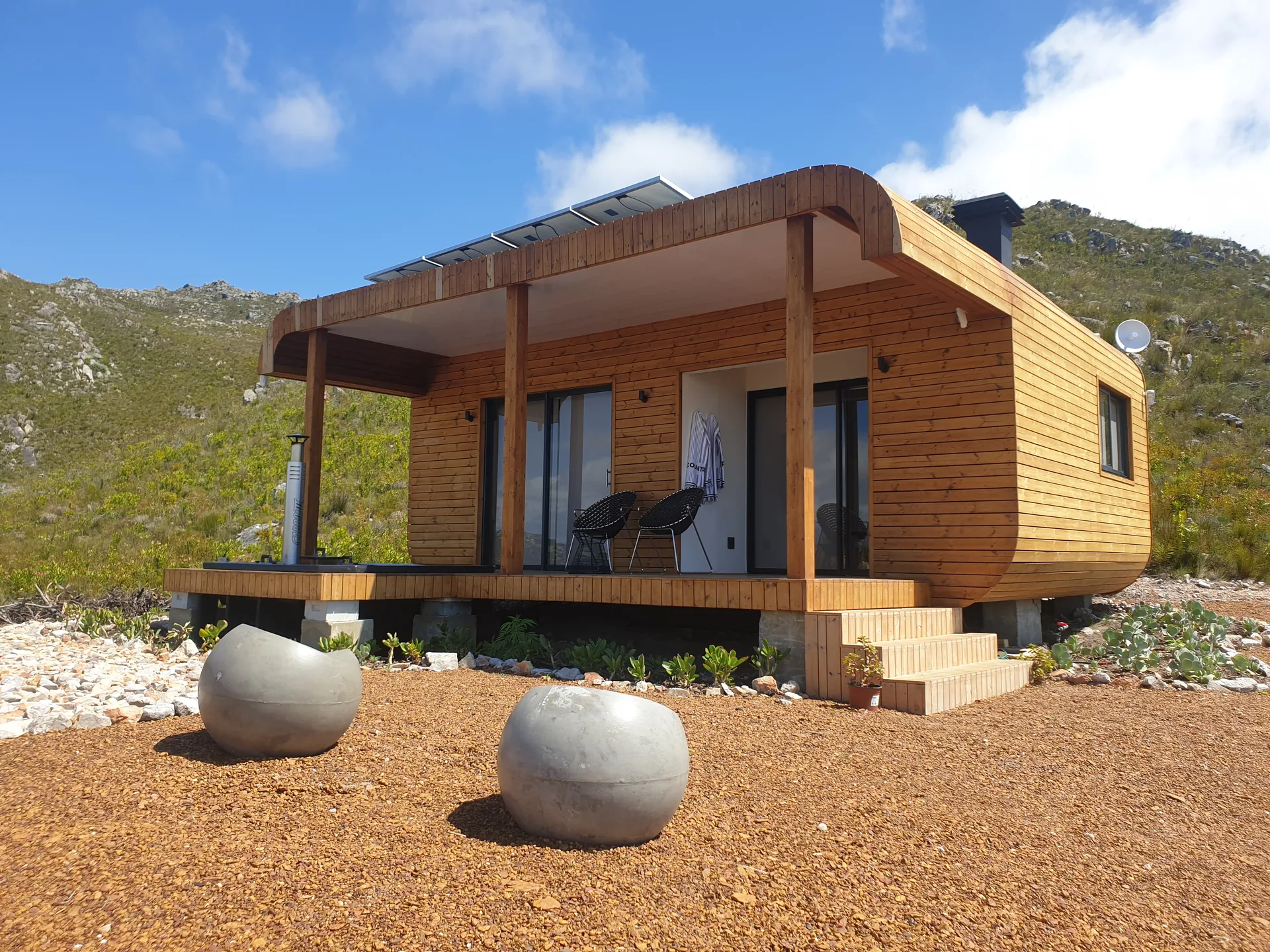 One-bedroom container house clad in timber, with a covered veranda and raised gravel setting below a mountain slope