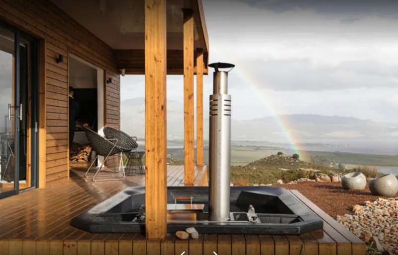 Container house deck with a wood-fired hot tub beneath the veranda and a rainbow over the surrounding landscape