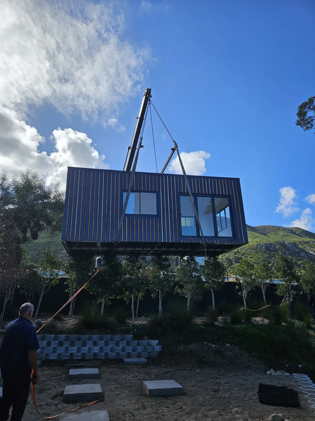 Dark clad modular unit suspended from a crane above a prepared site, with a worker guiding it by rope