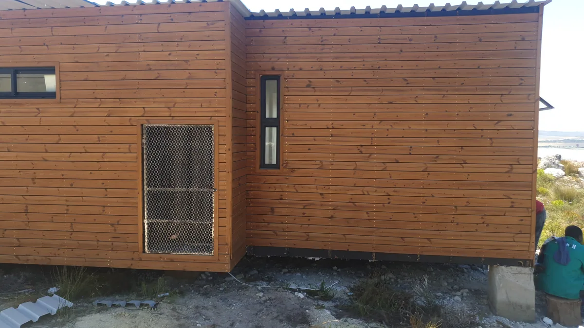 Timber-clad modular home exterior with a security-screened window, with a person partly visible at the edge of the frame
