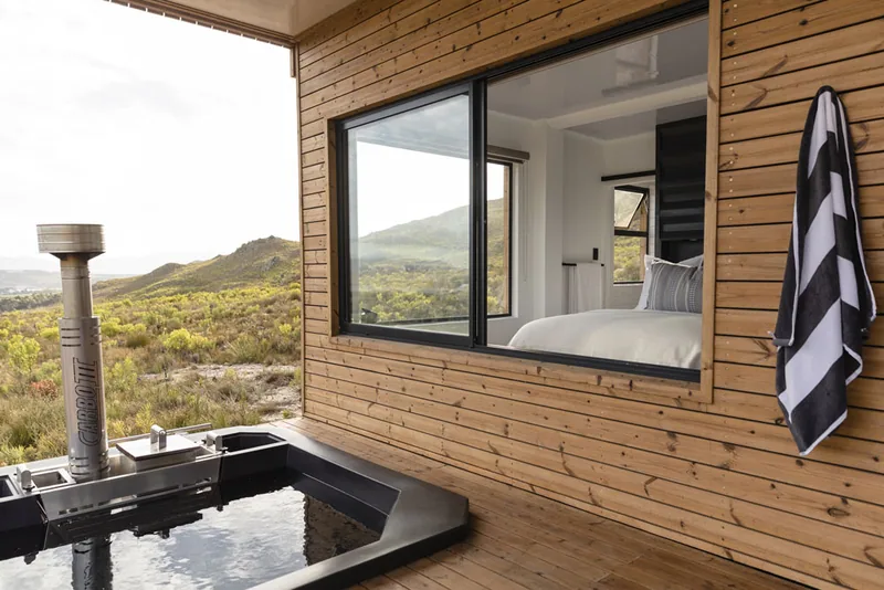 Outdoor bath on a deck beside a timber-clad home, with a towel hanging by the window and mountains behind