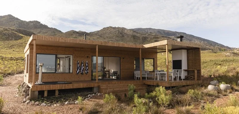 Timber-clad modular home with a covered veranda and outdoor furniture, set in open veld with mountains behind