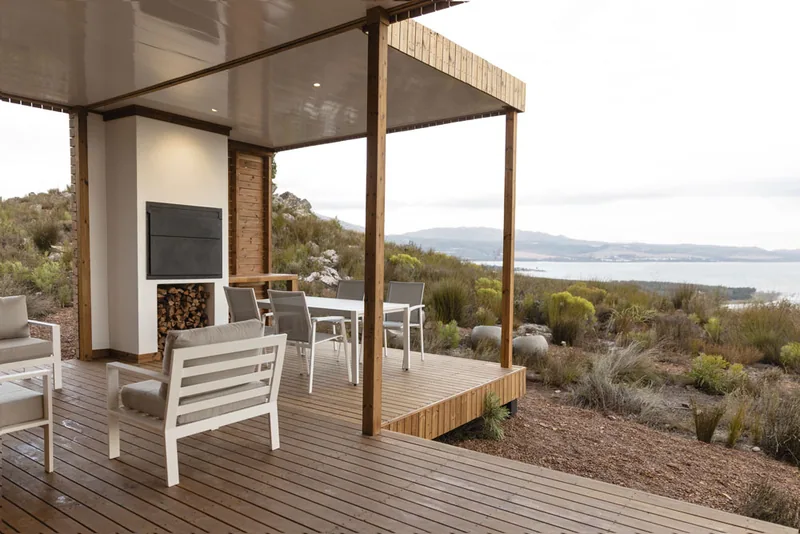 Covered veranda with outdoor seating, a built-in fireplace and timber posts, looking across scrub to a lagoon