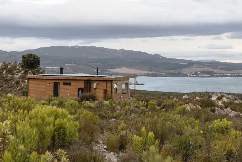 Timber-clad modular home set in coastal fynbos with a lagoon and mountain range in the background