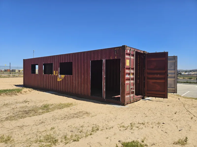 Two red containers positioned on a sandy site with door and window openings cut into the side walls