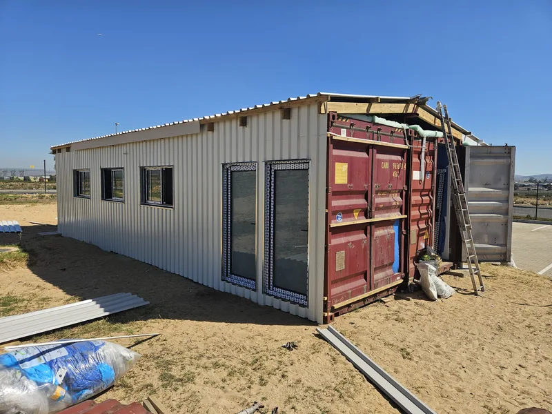 Container building under construction, half clad in white IBR sheeting with a red container still exposed and a water tank alongside