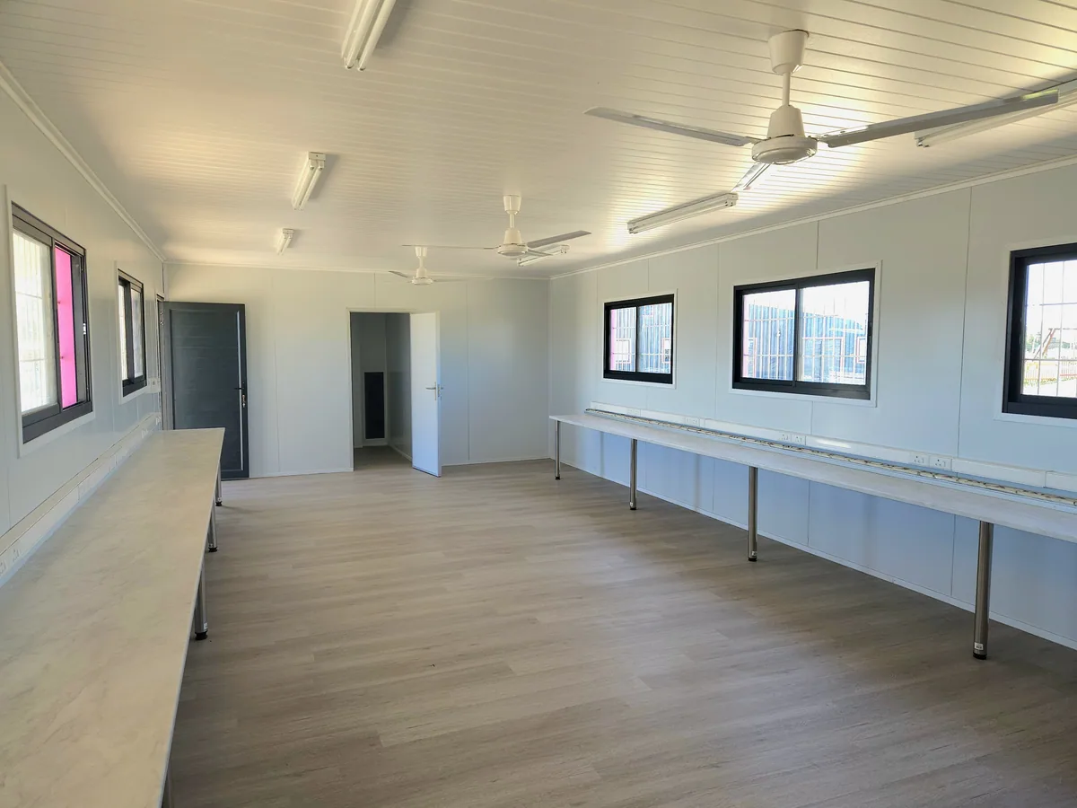 Long classroom interior with a continuous white worktop on steel legs, ceiling fans and windows along one wall