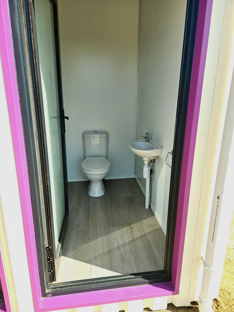 Bathroom inside a container classroom with a white toilet and basin, framed by a bright pink door surround