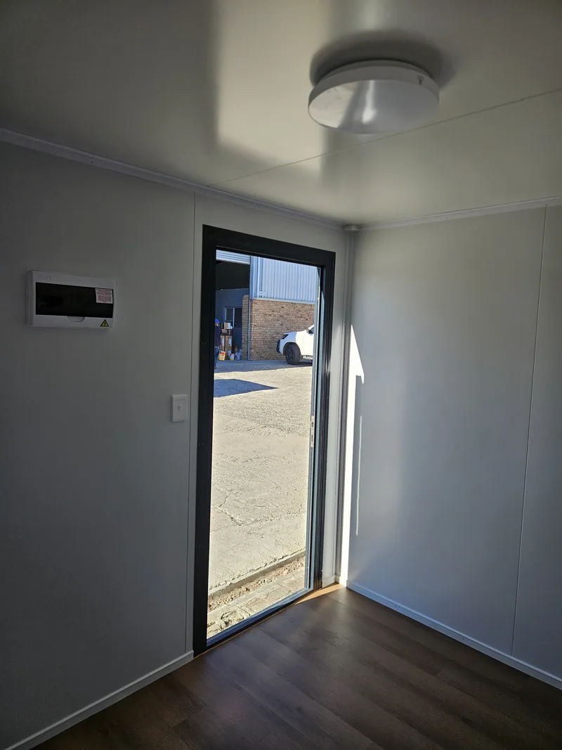 Office interior looking through an open black door to the yard outside, ceiling light overhead