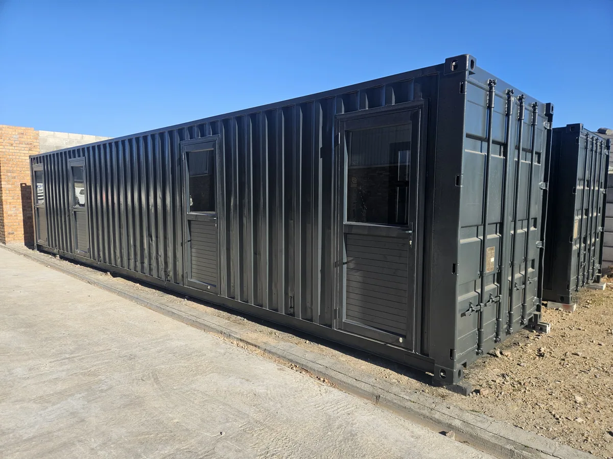 Long black 12m container office with windows and a louvred vent, alongside a brick building; container markings visible