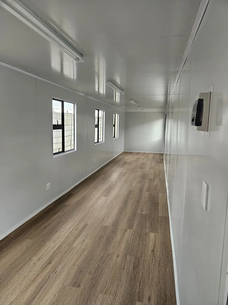 Empty open-plan container office interior with white walls, wood-look flooring and windows along one side