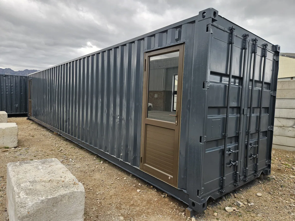 Long dark container office with a timber-framed glazed door, standing beside a concrete block