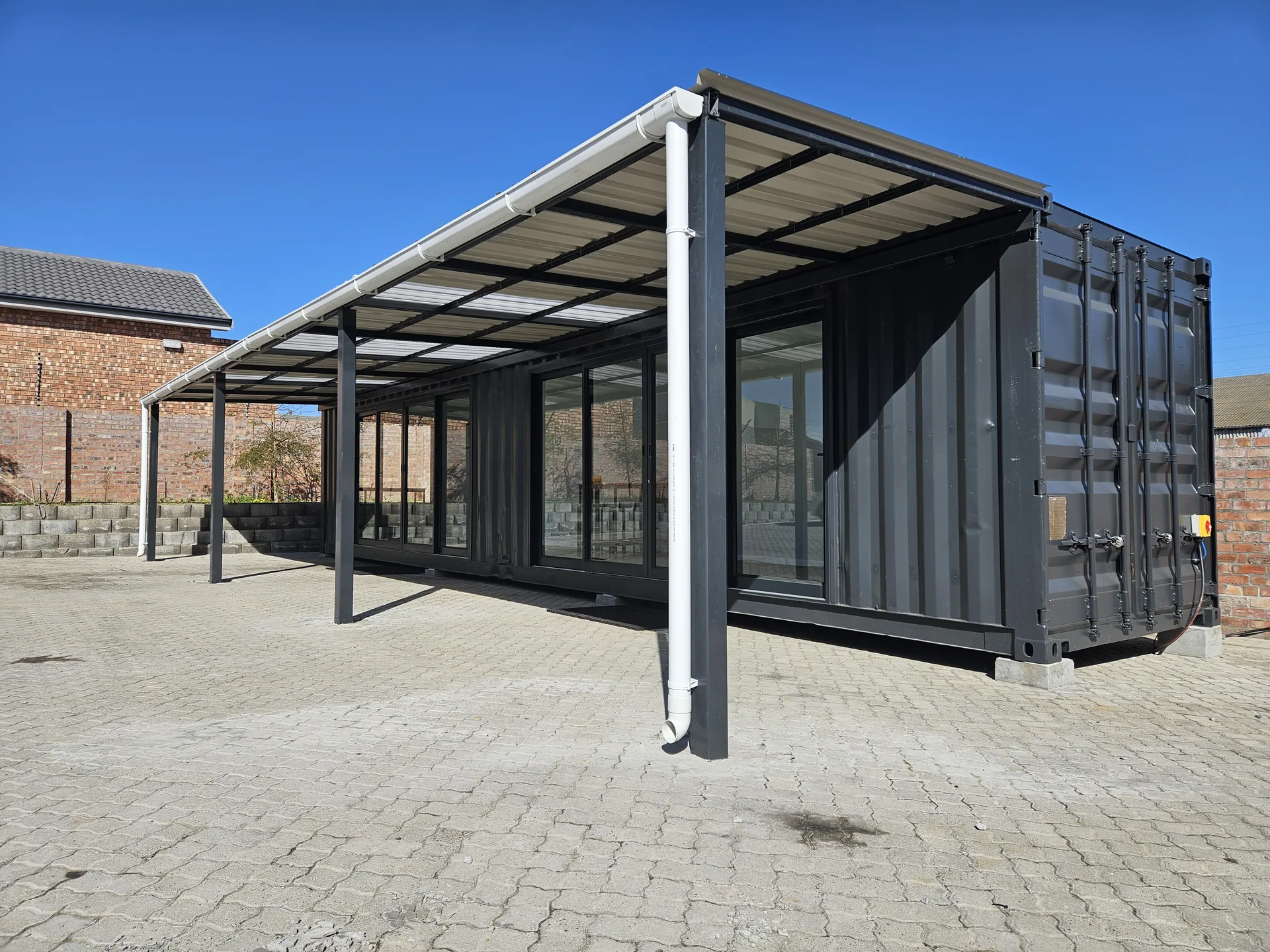 Container office with full-height glazing and a large steel and polycarbonate veranda canopy over a paved courtyard