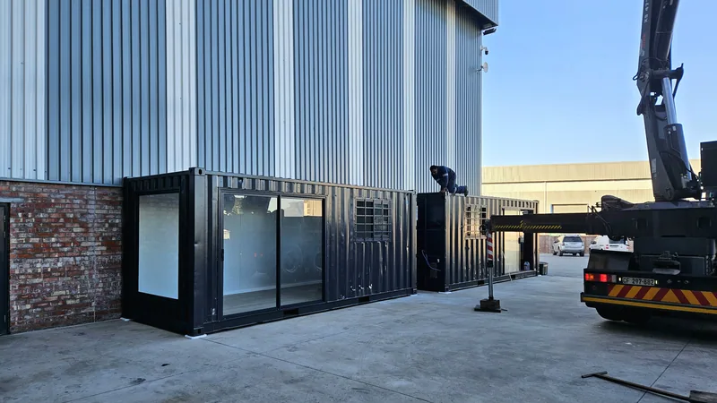 Container office being positioned beside a brick and steel building, with a truck-mounted crane and vehicles in the yard