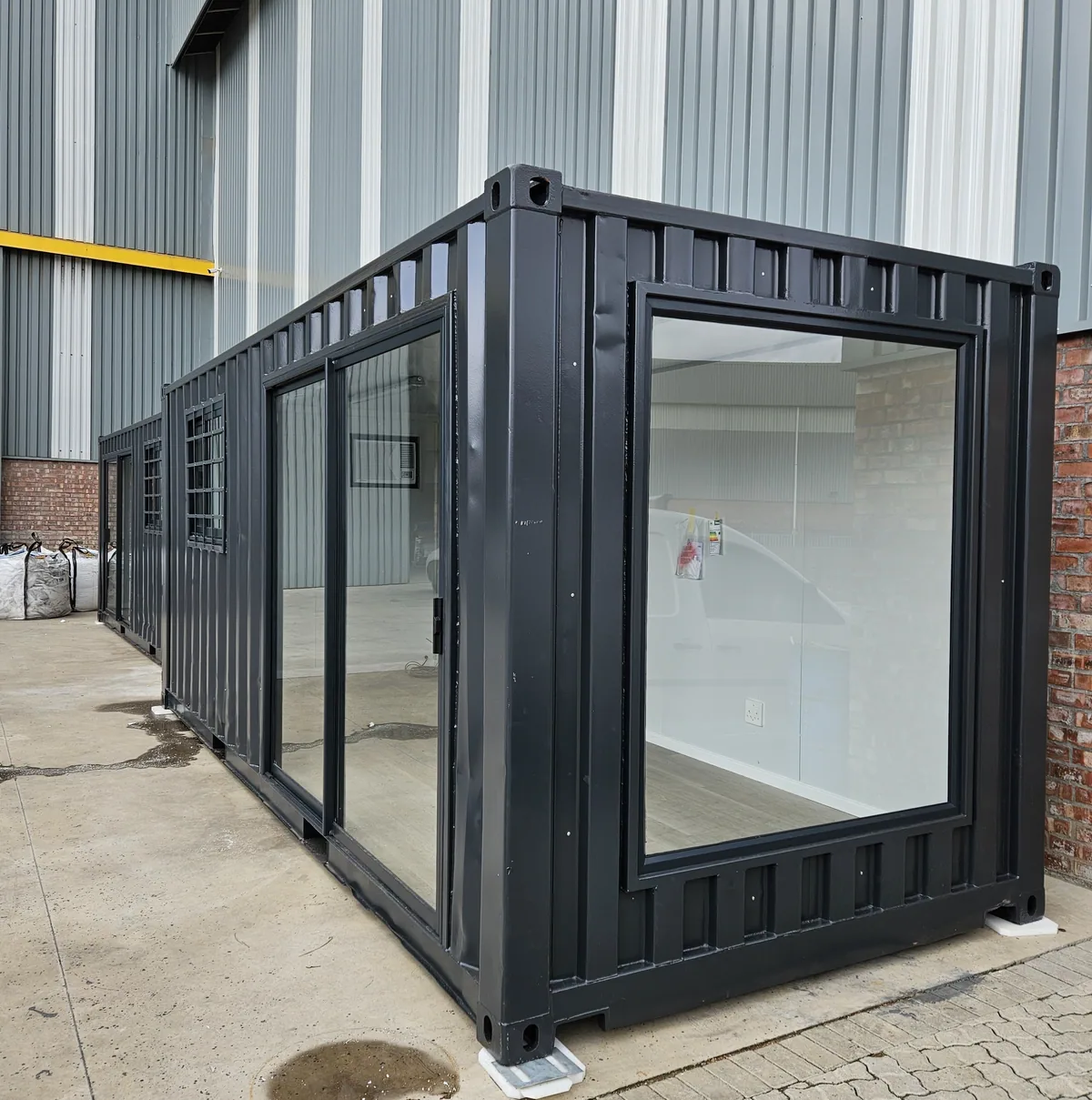 Black 6m container office with a large glazed corner window, standing on a paved yard