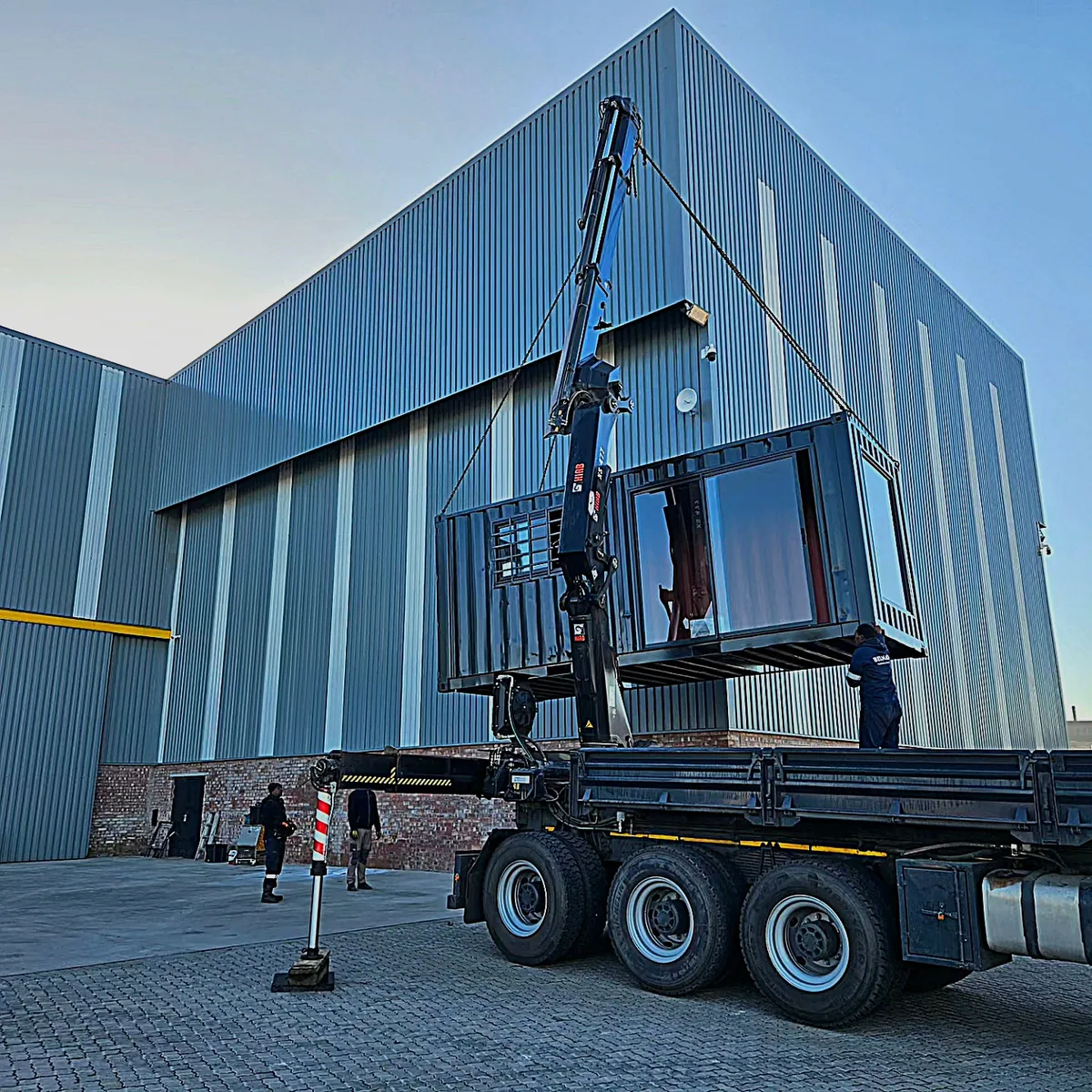 Truck-mounted crane lifting a container office off a flatbed truck beside a blue-clad industrial building, with workers on the ground