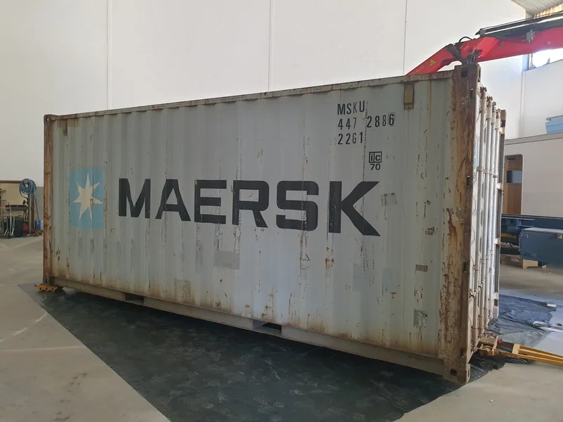 Weathered white shipping container with third-party shipping line branding, being moved inside a workshop