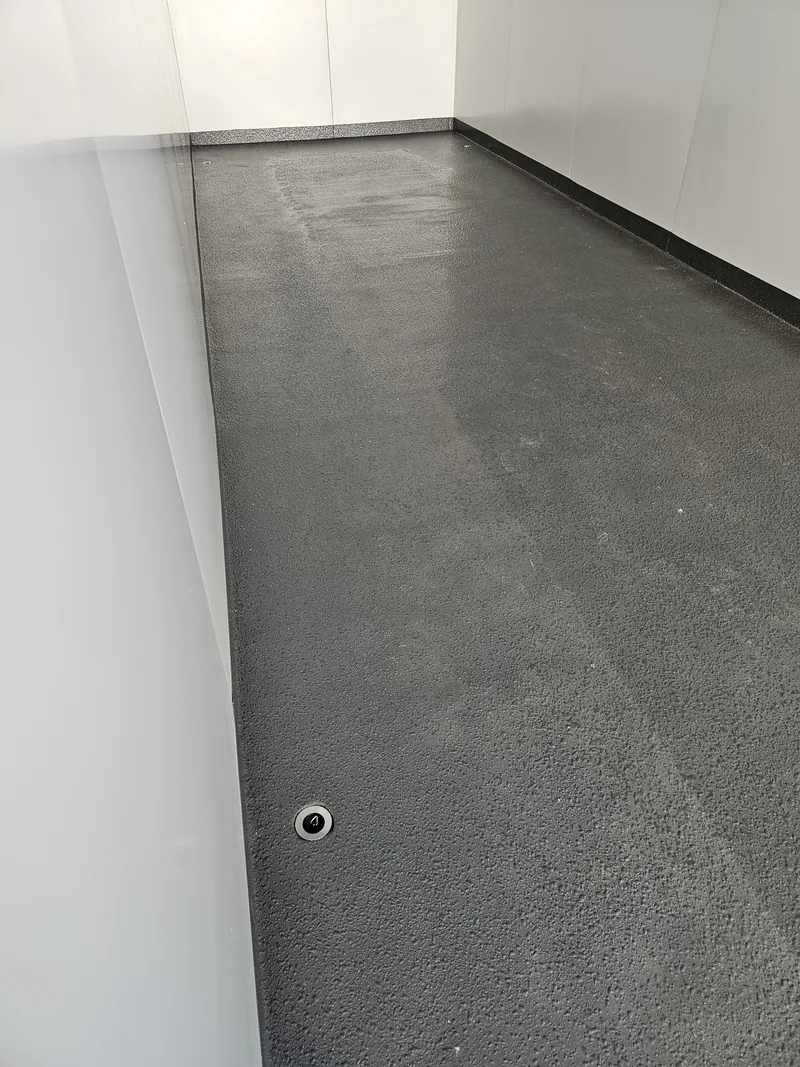 Close view of a black epoxy floor coating meeting a white wall inside a container, with a floor drain fitted
