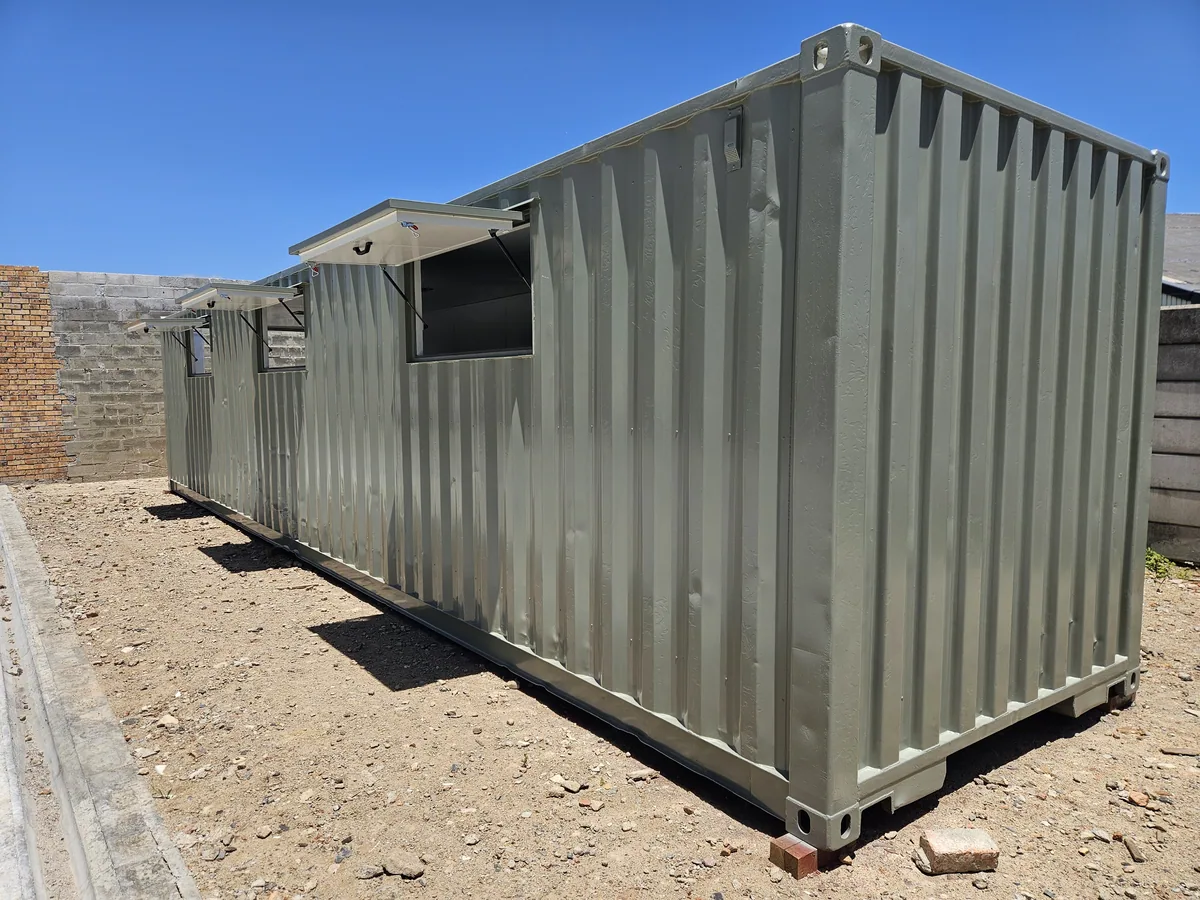 Pale green container kitchen unit with a serving hatch, standing on a paved site beside a brick wall