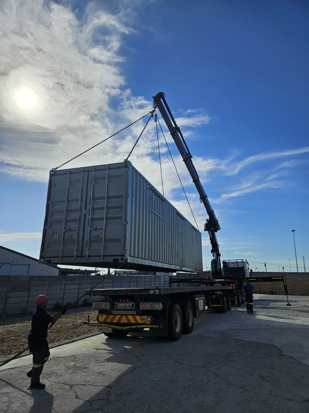 Crane lifting a grey container from a flatbed truck under a bright sky, with a worker in a hard hat guiding from the ground