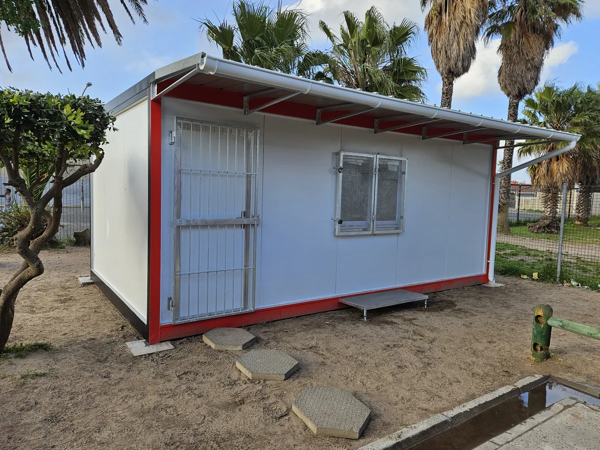 White modular kitchen unit with red trim and a steel-framed security door, standing on a sandy yard under palm trees