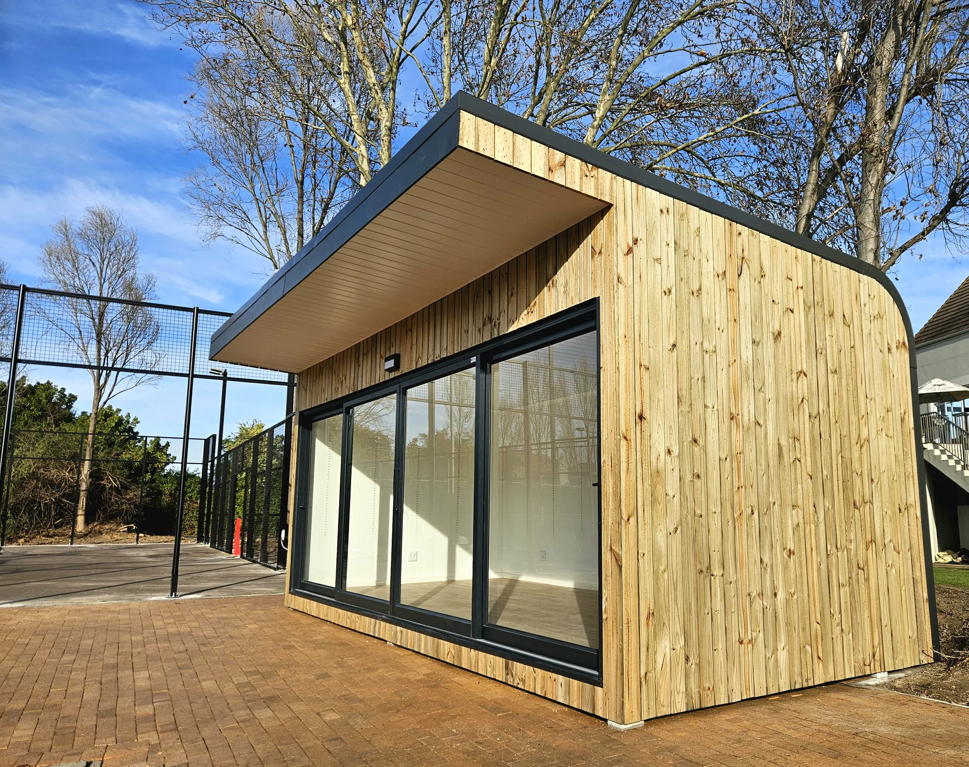 Timber-clad modular pro shop with an angled roof and full-height sliding glass doors on a paved court surround