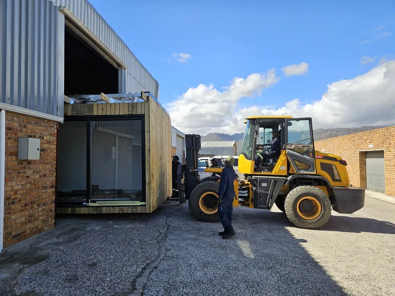 Modular unit being manoeuvred beside a brick building by a front-end loader, with a worker alongside