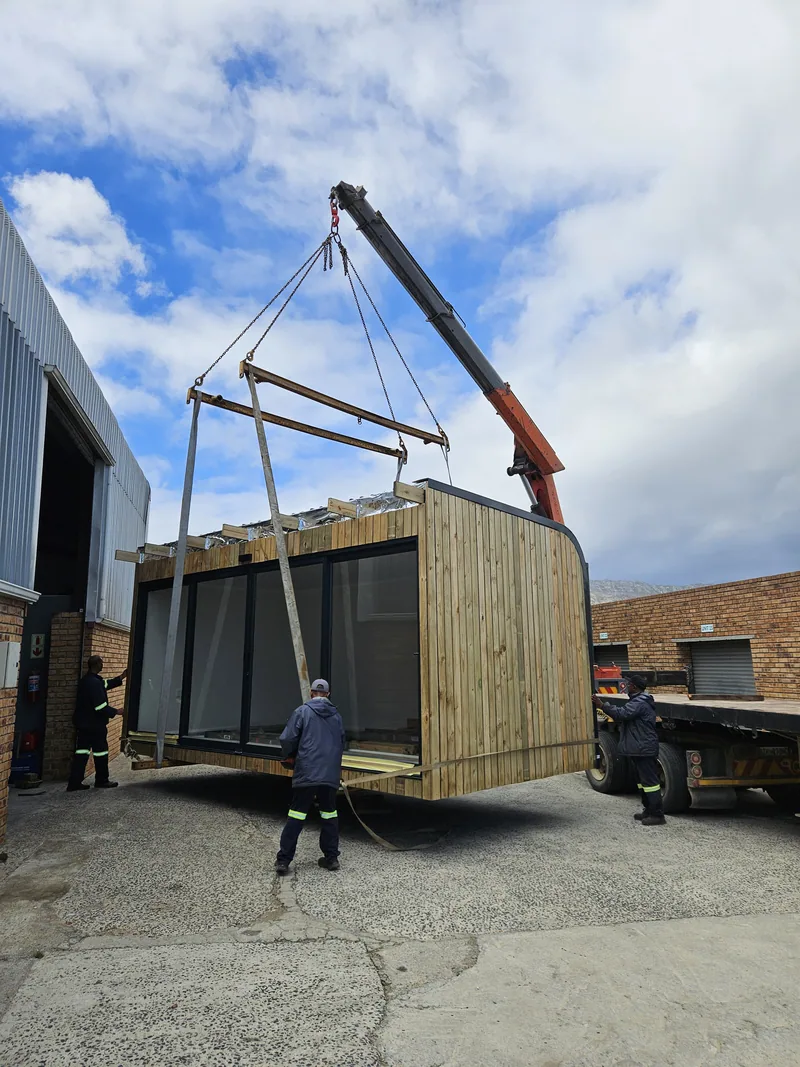Timber-clad modular unit lifted by a crane outside a workshop, with a worker steadying it from below