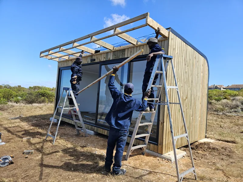 Workers on ladders fixing roof purlins to a timber-clad pod on a coastal site