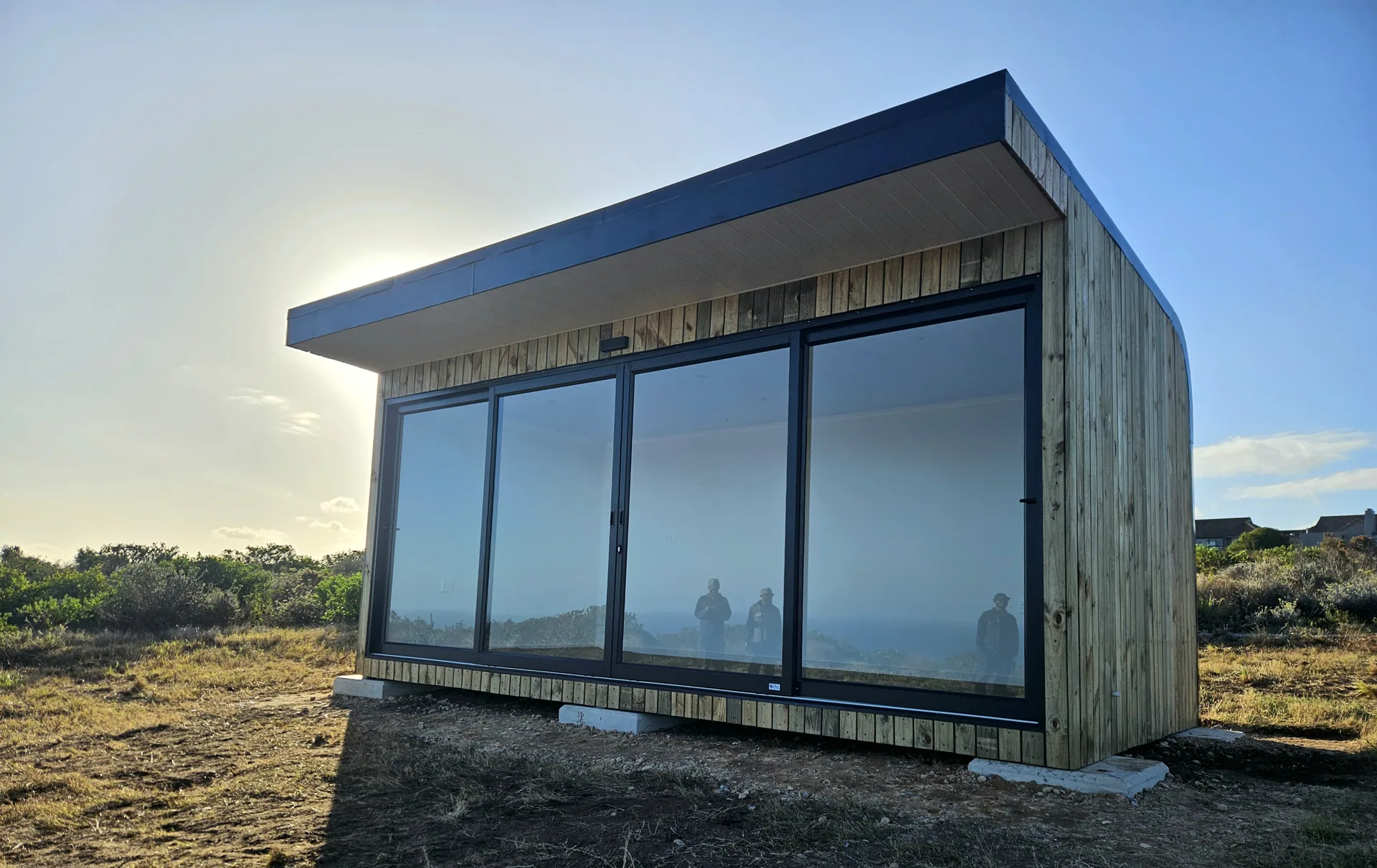 Timber-clad glazed pod at dusk on a coastal site, with warm light on the cladding