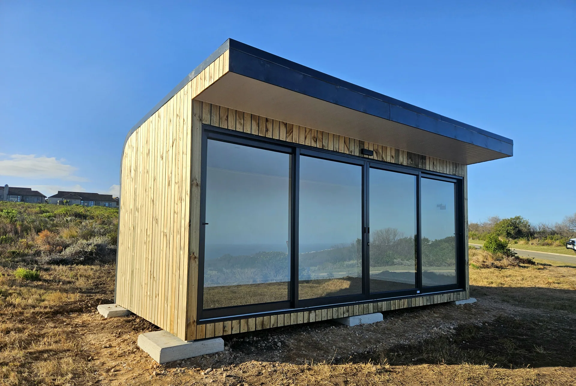 Timber-clad sales pod with a wraparound glazed corner, standing on concrete pads in coastal scrub