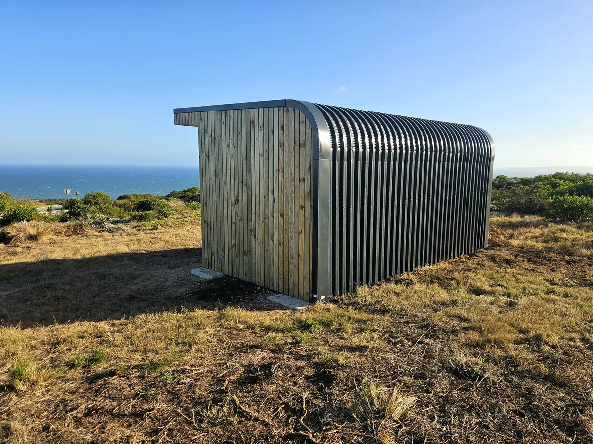 Side view of a pod with curved corrugated steel cladding and timber ends, on a hillside above the sea