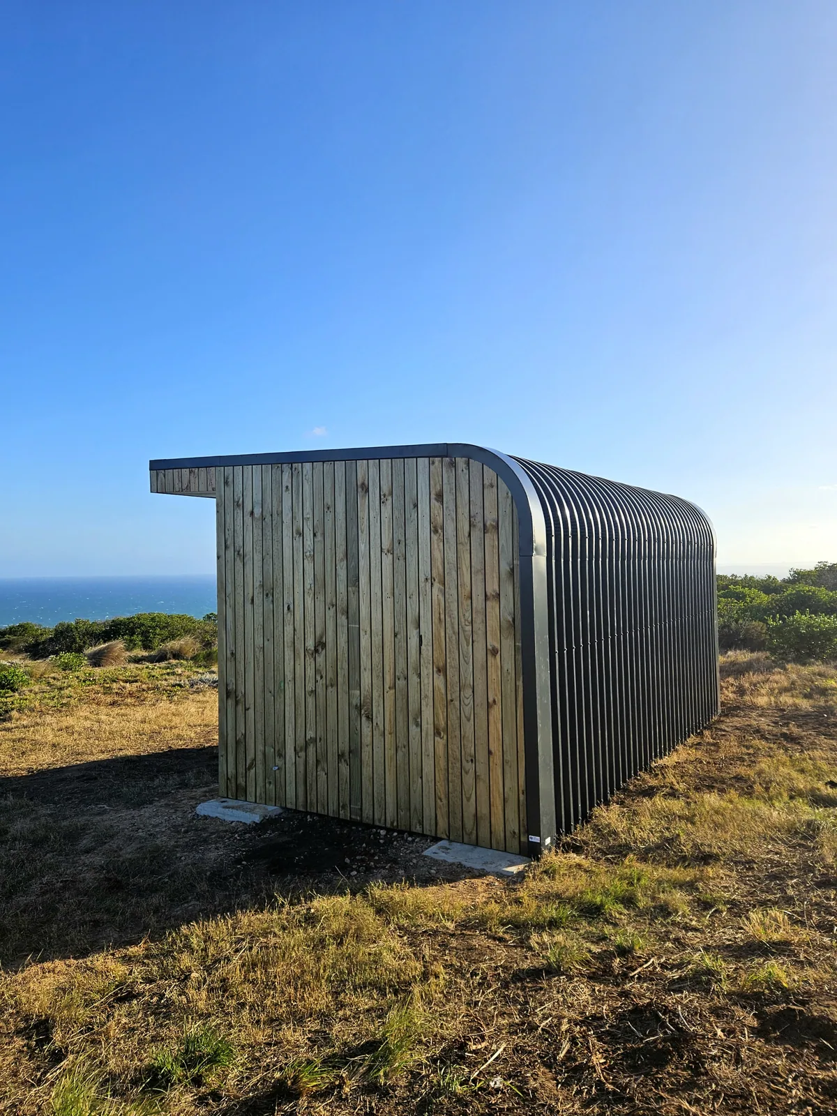 Corrugated steel and timber-clad pod standing on a grassy coastal slope