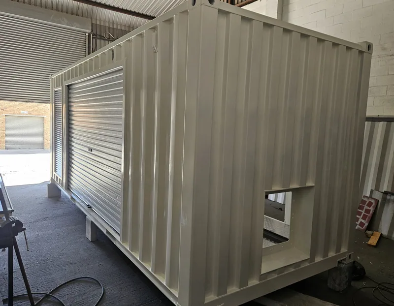 Container with a roller shutter door and service hatch, open at the end and standing on a workshop floor