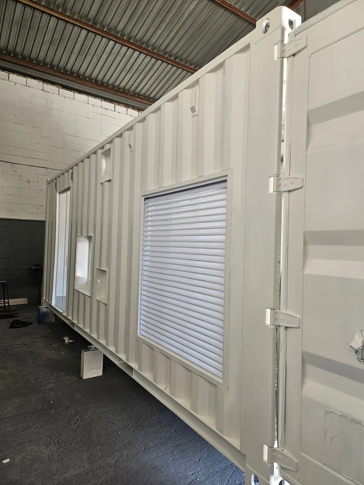 White container fitted with a roller shutter and window, standing on levelling blocks in a workshop