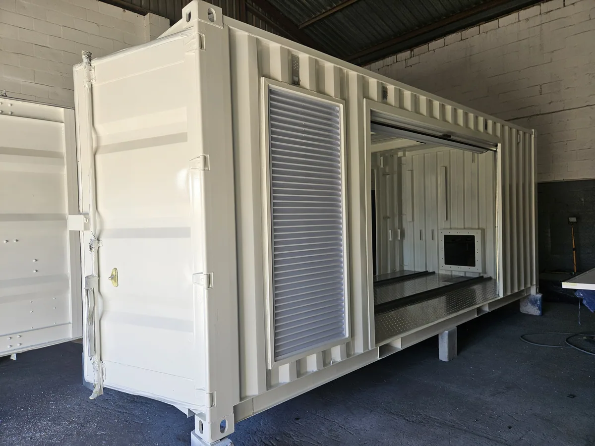 White containers with roller shutters and service hatches lined up inside a fabrication workshop