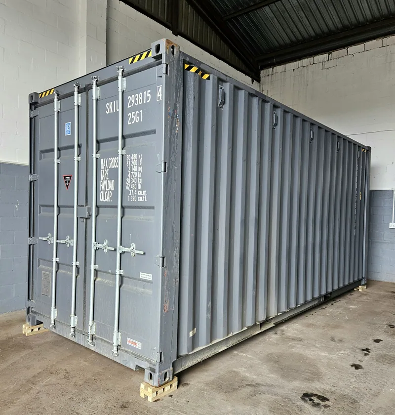 Blue-grey shipping container on timber blocks inside a workshop, with container markings on the side