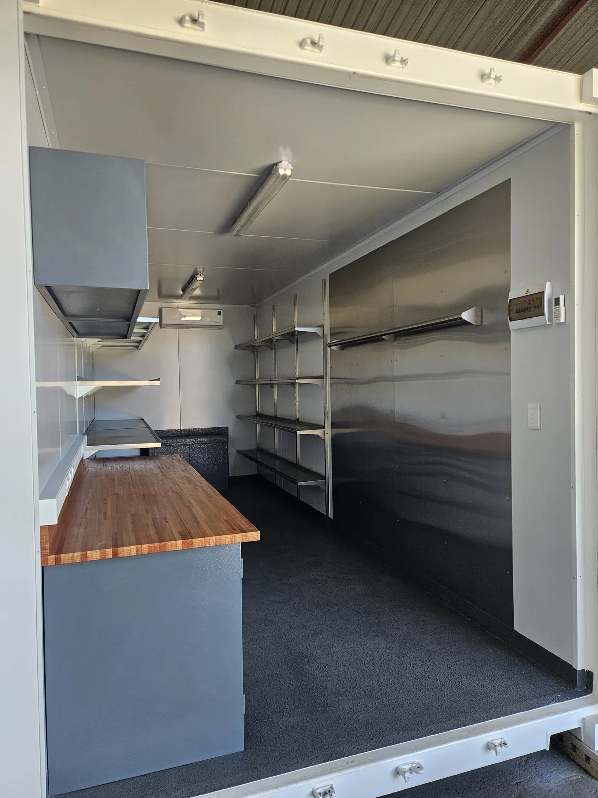 Container workshop interior with the side doors open, showing a timber bench, wall cabinet and shelving